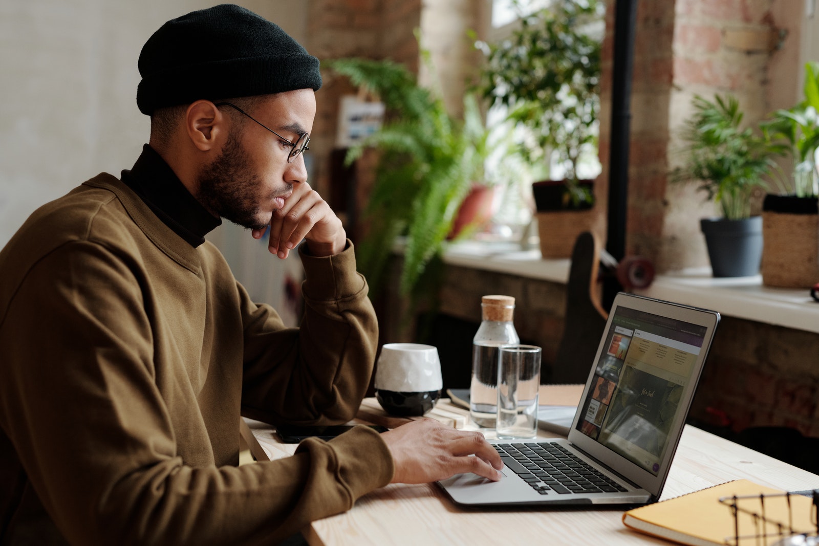 Man in Brown Coat and Blue Knit Cap Sitting by the Table Using Macbook Pro