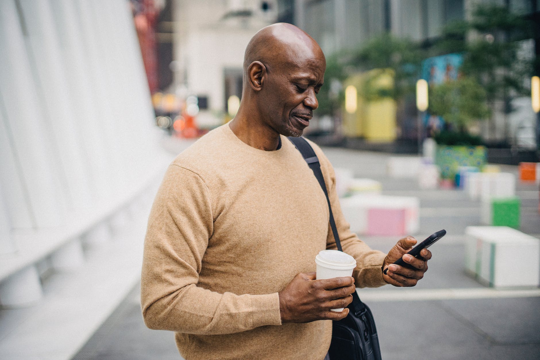 pensive mature black male with takeaway coffee using smartphone on street