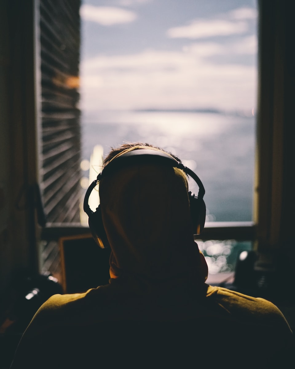tilt-shift lens photography of person wearing headphones facing body of water through window of a dark room